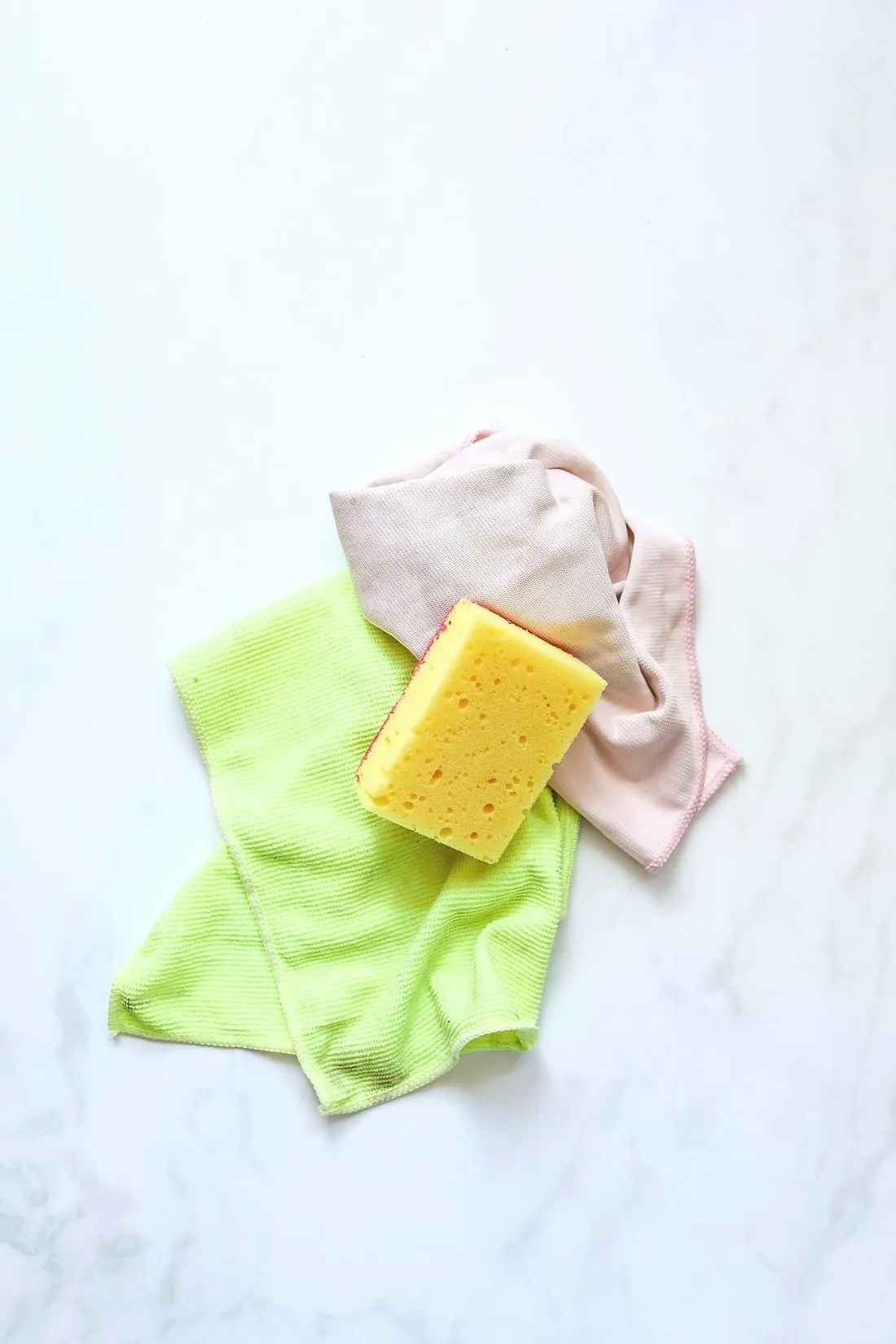 A yellow sponge, a lime green microfibre cloth and a pink cloth on white marble
