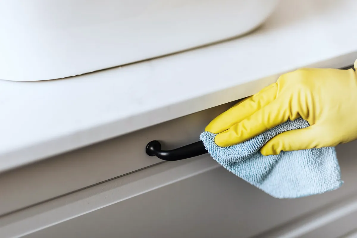 A hand in a yellow rubber glove wiping a white cupboard handle with a blue cloth