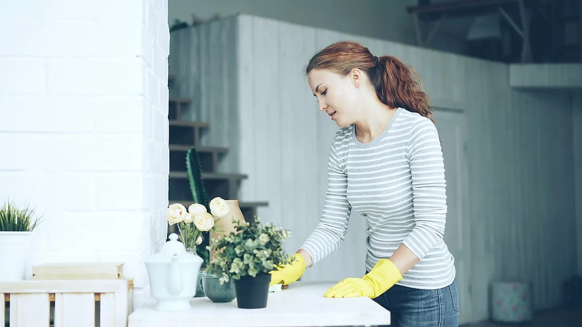 A woman in yellow rubber gloves wiping a white table next to a vase of flowers in a bright room