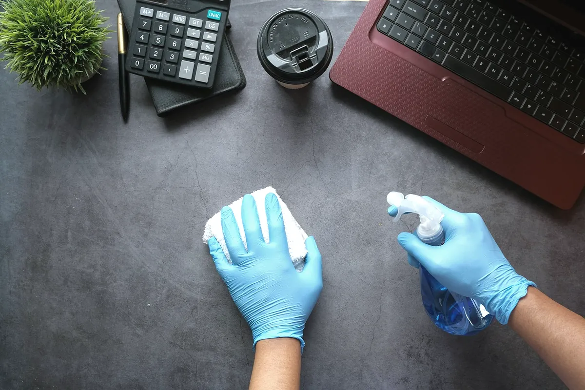 Hands in blue gloves wiping a dark desk next to a laptop, a calculator and a coffee cup