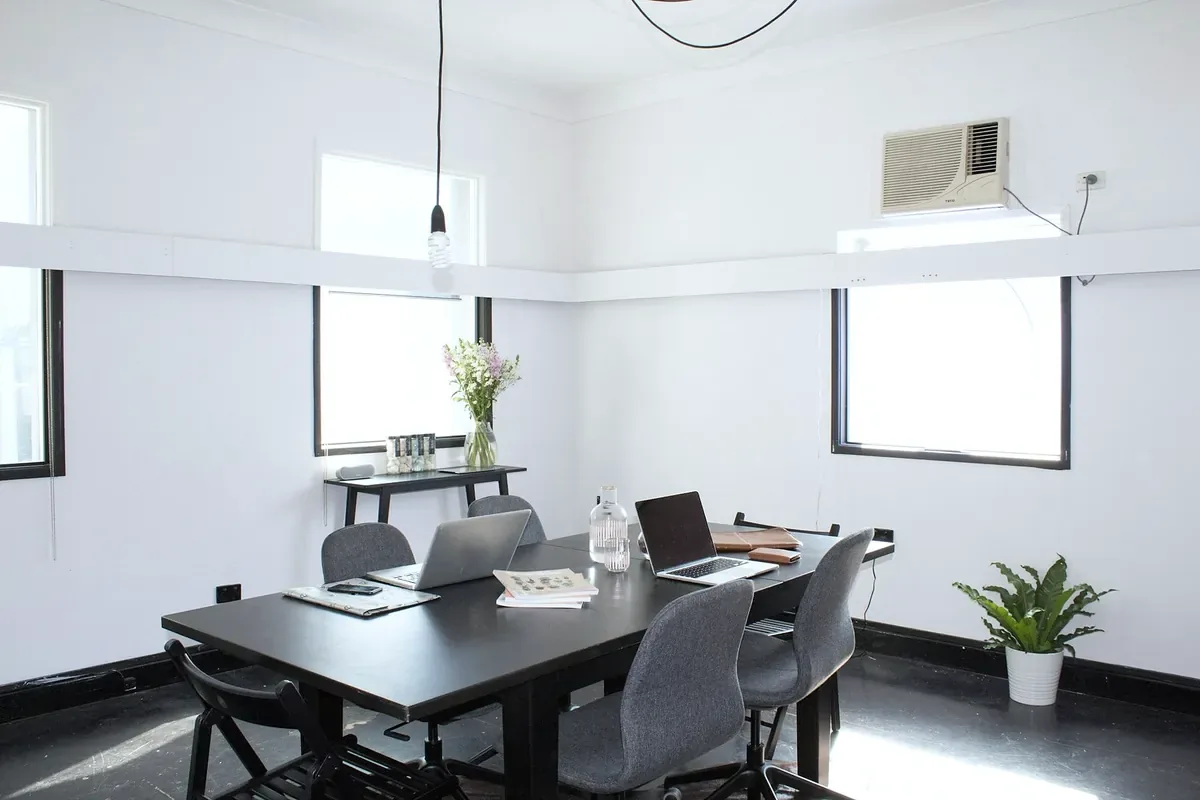 A small white office with a dark meeting table, grey chairs, laptops and a plant