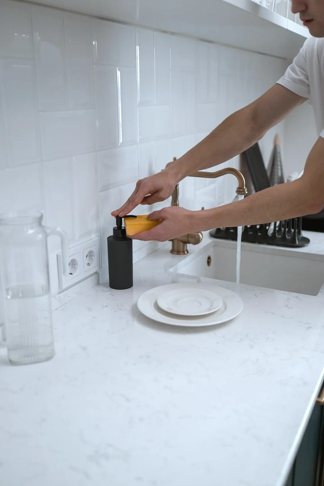 A cleaner pressing a soap dispenser over a sponge at a white kitchen sink