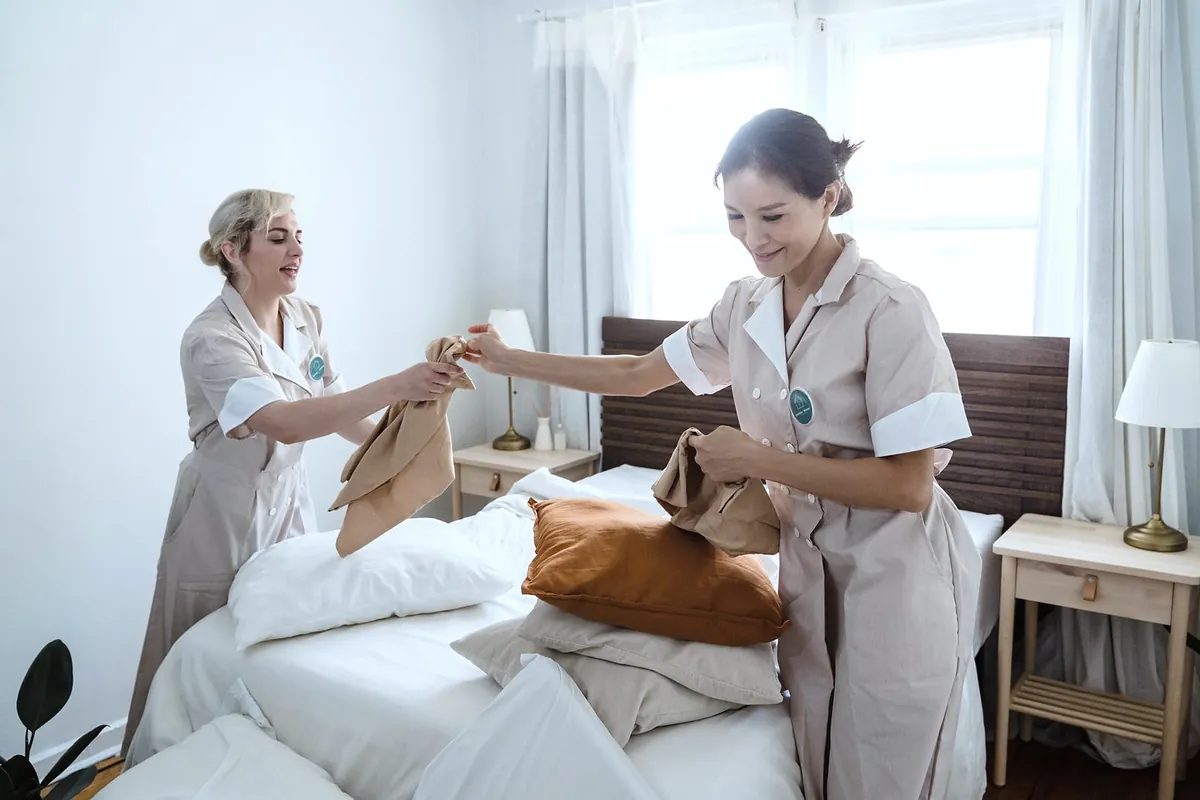 Two cleaners in light uniforms making up a bed with white linen in a bright bedroom