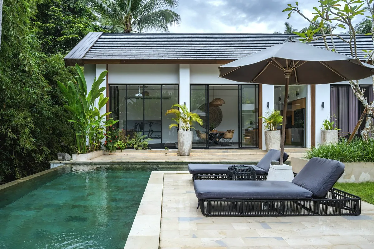 A single storey holiday villa with a long pool, sun loungers and a parasol in a tropical garden