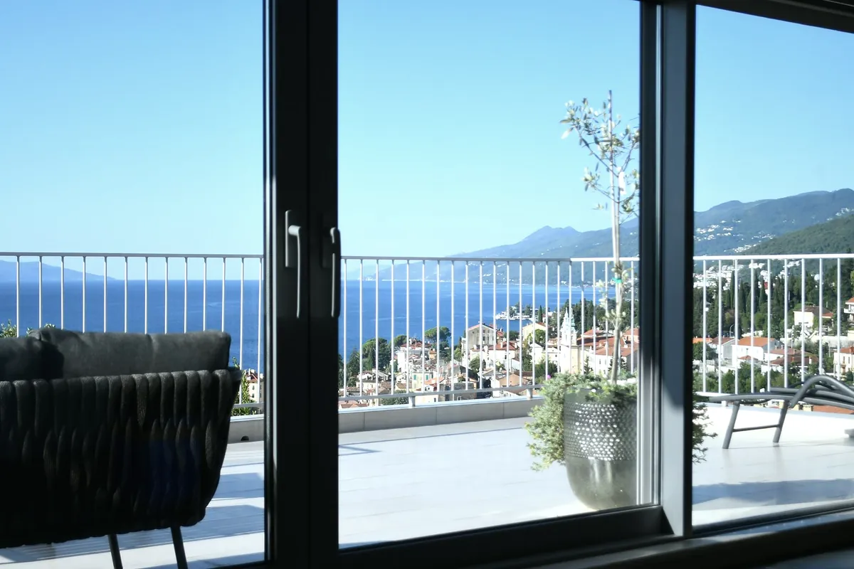 A villa terrace seen through open glass doors, with the sea and hills beyond