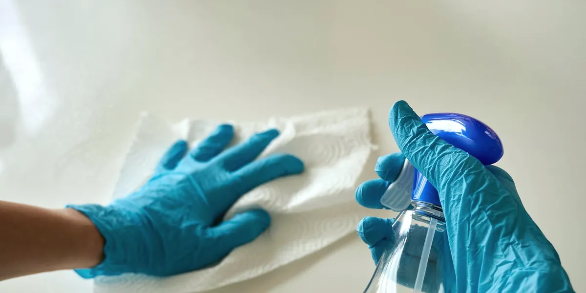 Hands in blue gloves wiping a white worktop with paper towel and a spray bottle