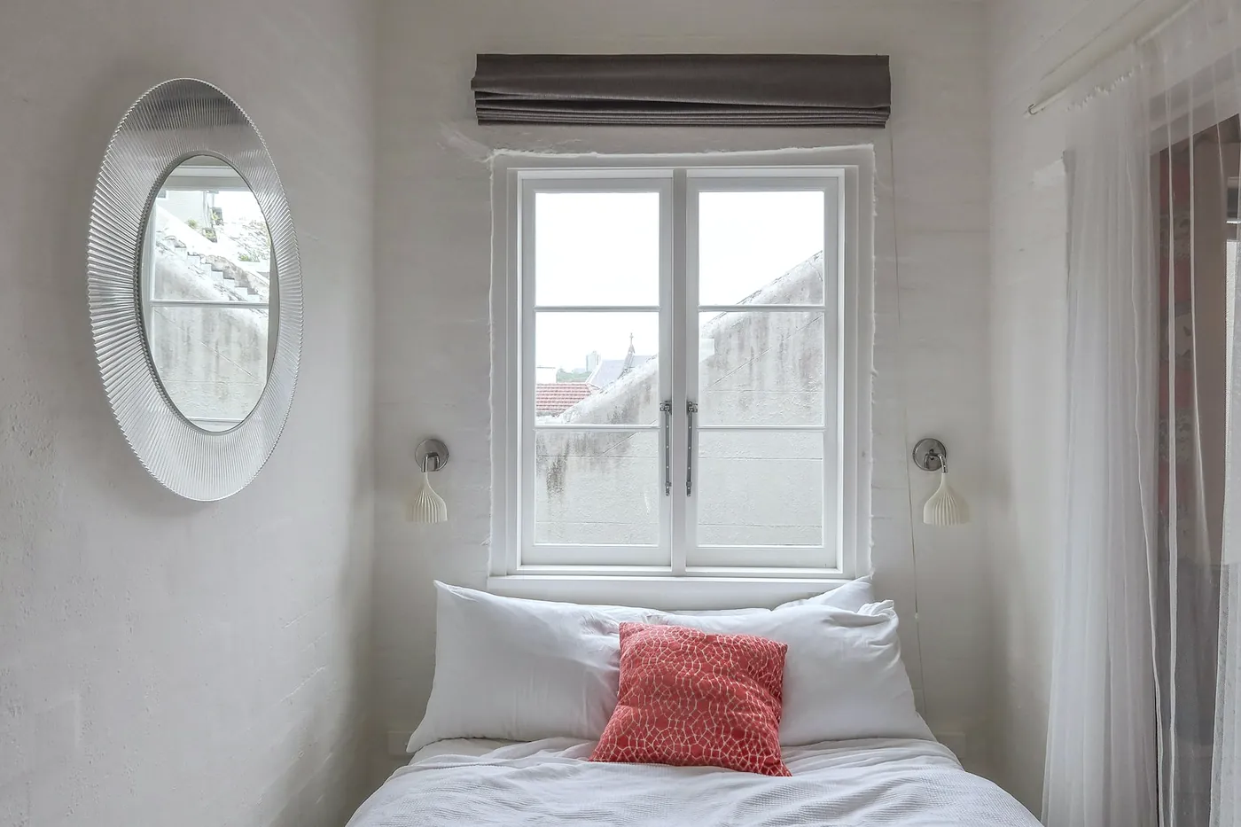 Small white bedroom with a double bed under a window, a round mirror and a coral cushion