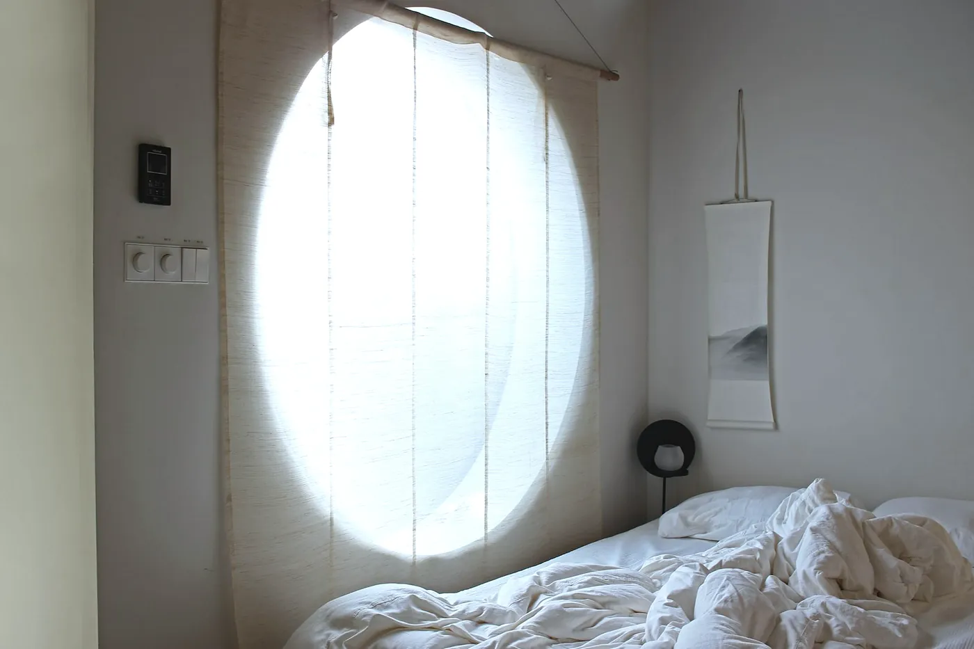 Bedroom with a large round window covered by a linen blind and an unmade white bed