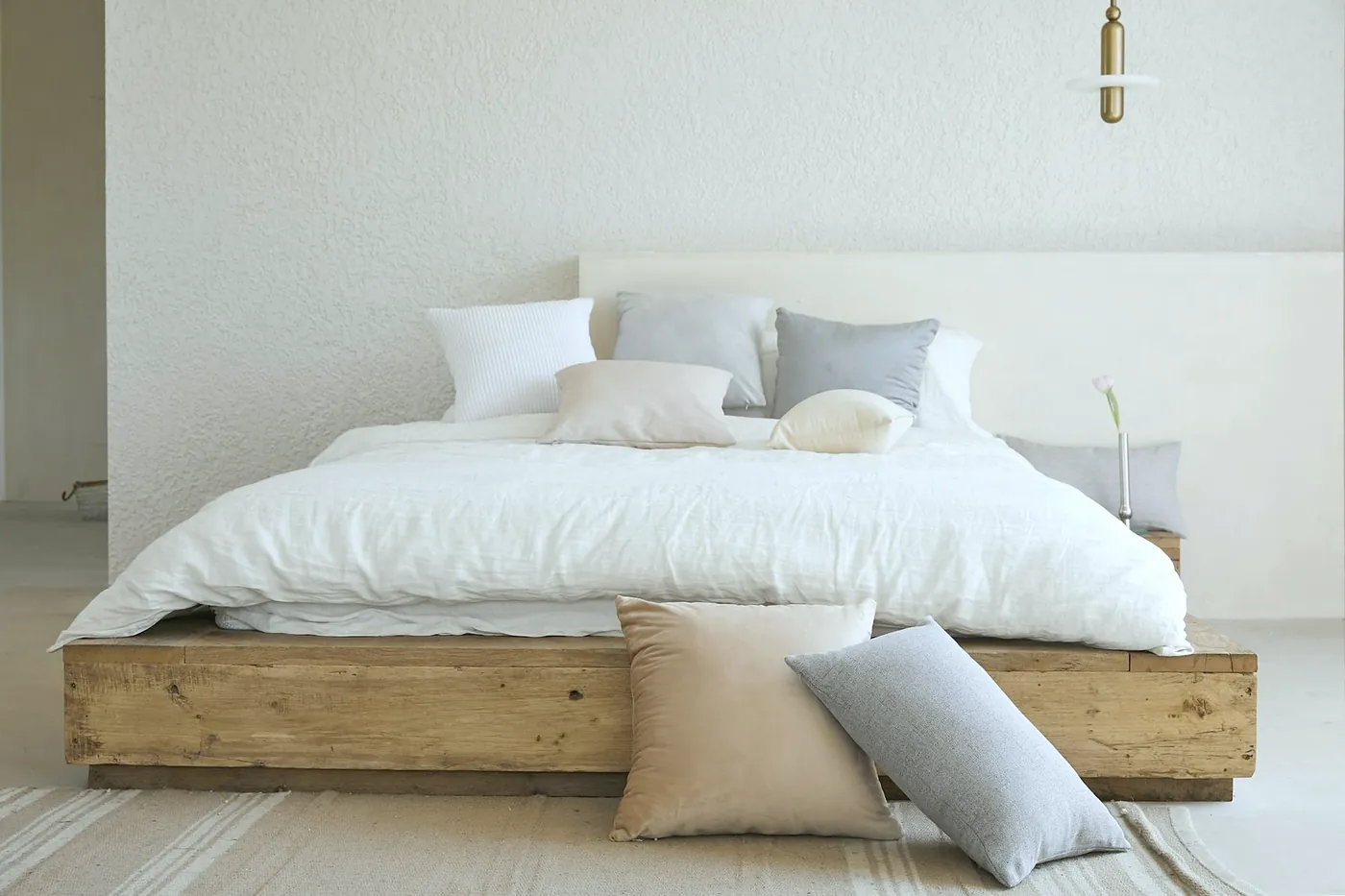 Low oak bed with white linen and grey and sand coloured cushions against a white wall