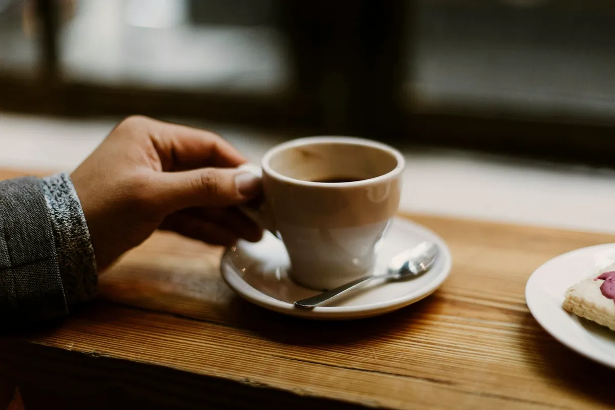 A hand lifting a cup of coffee from its saucer on a wooden table by the window, a slice of cake on a plate beside it