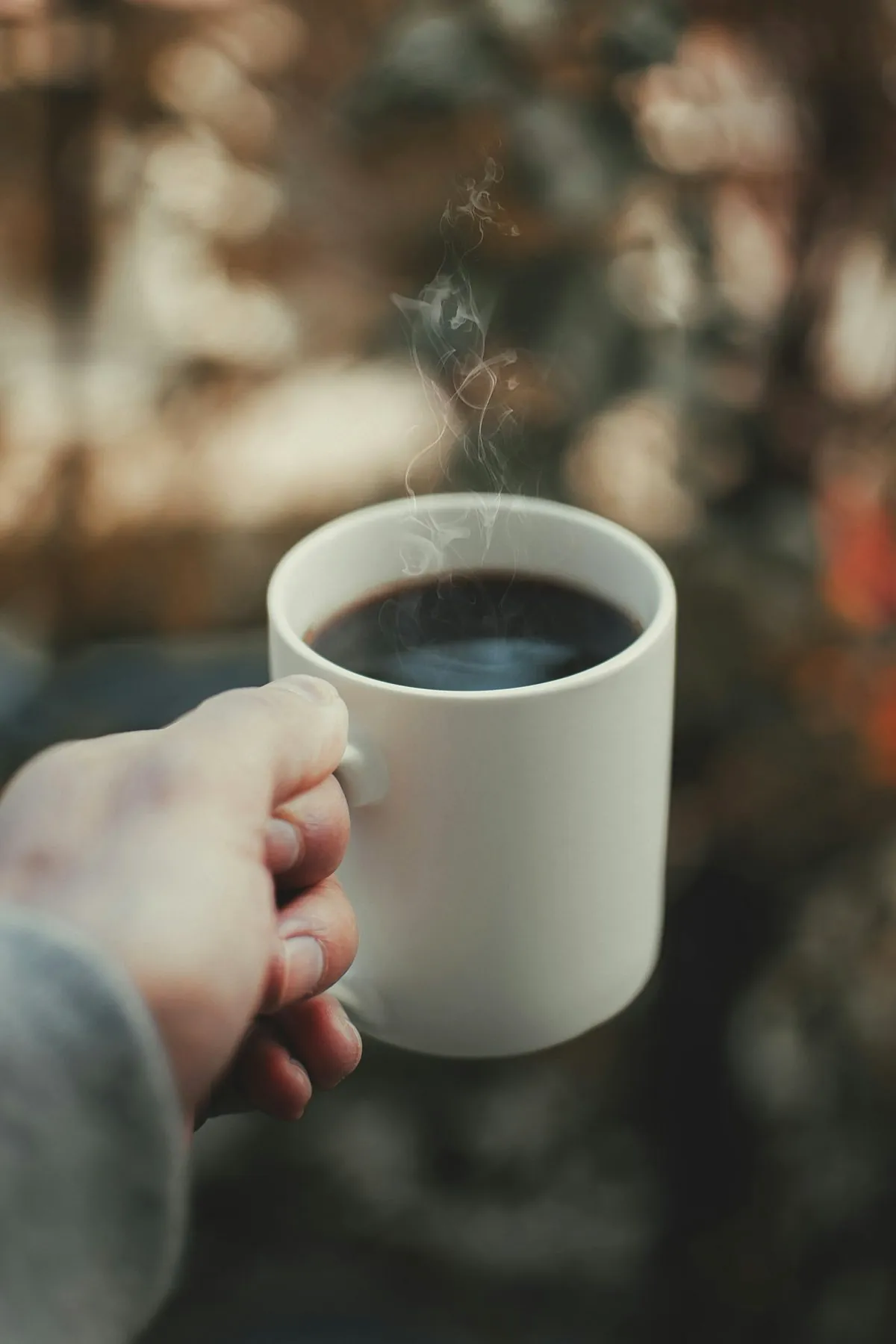 A hand holding a white mug of black filter coffee, steam rising from it in the morning light