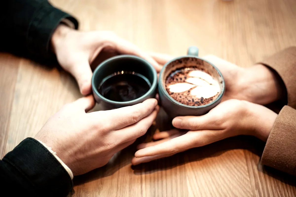 Two people holding their coffee cups together across a wooden table