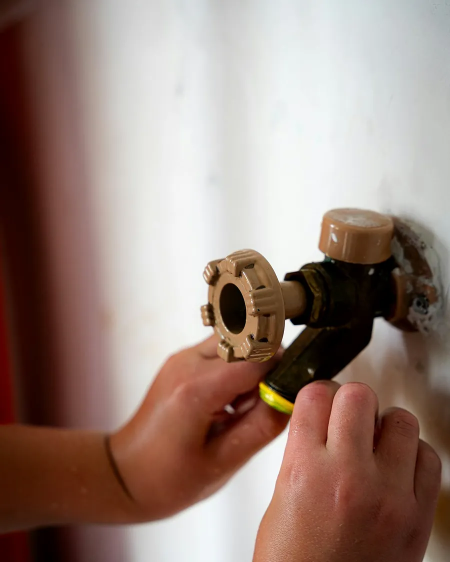 Two hands turning a brass stopcock where the water pipe comes out of a white wall