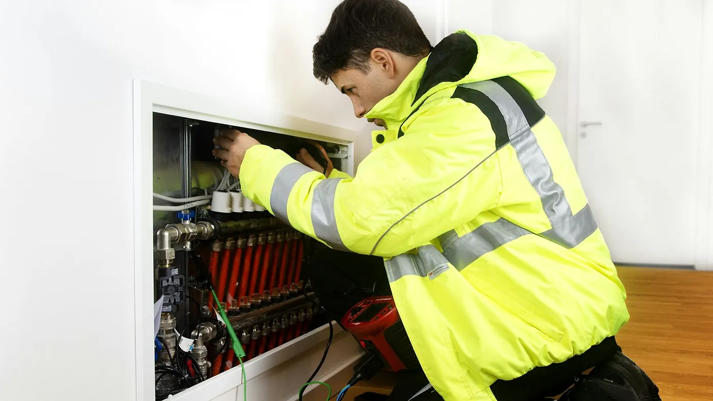 A plumber in a yellow high visibility jacket kneeling at an open underfloor heating manifold in a white hallway