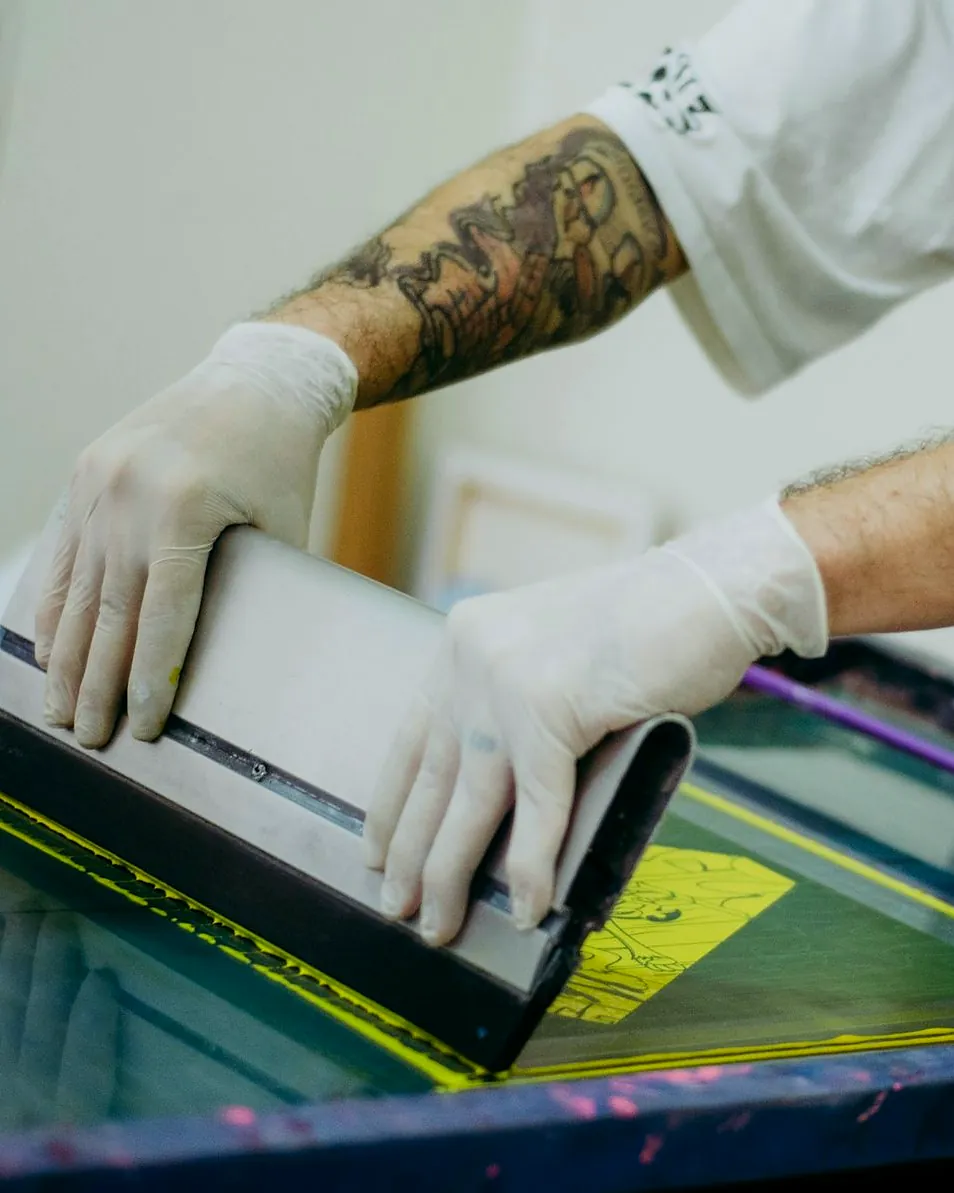 Gloved hands pulling a squeegee through yellow ink on a screen.
