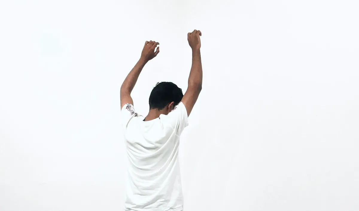 A man seen from behind against a white wall, raising both arms above his head