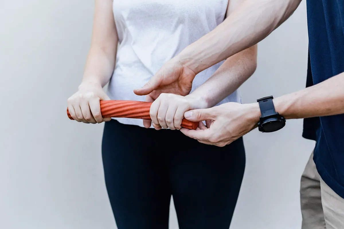 A woman twisting an orange flexible bar while a physiotherapist guides her wrists