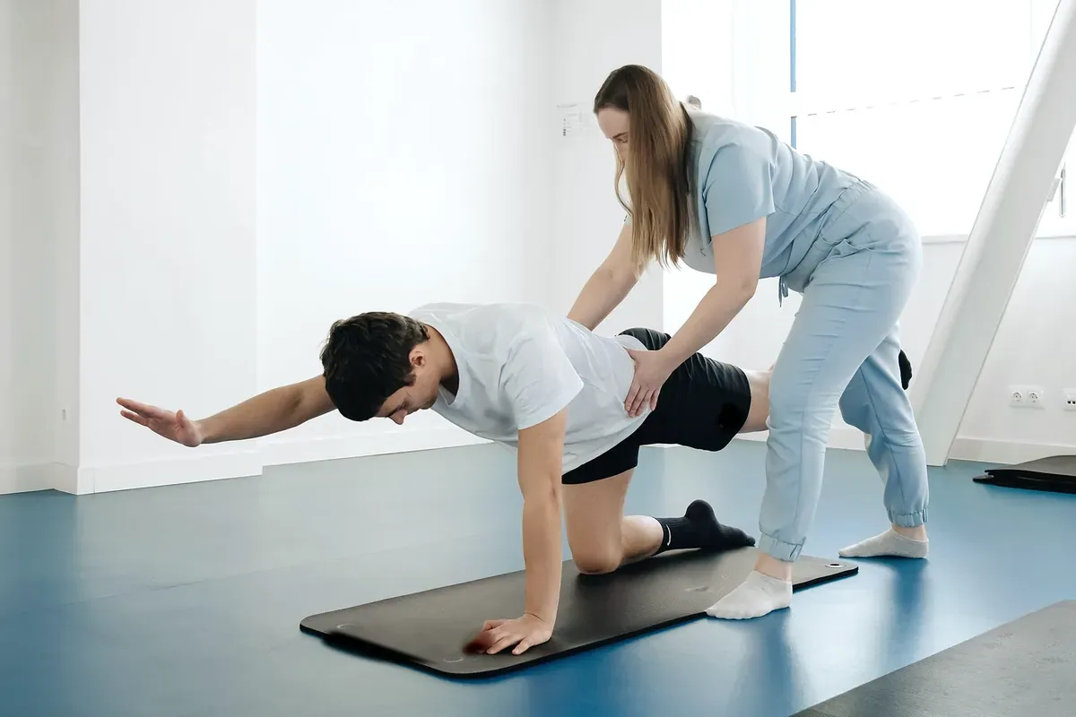 A physiotherapist steadying a man on all fours on an exercise mat as he reaches one arm forward