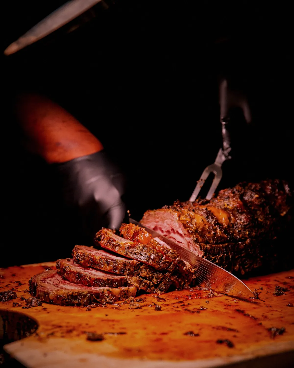A gloved hand carving slices from a roast on a wooden board