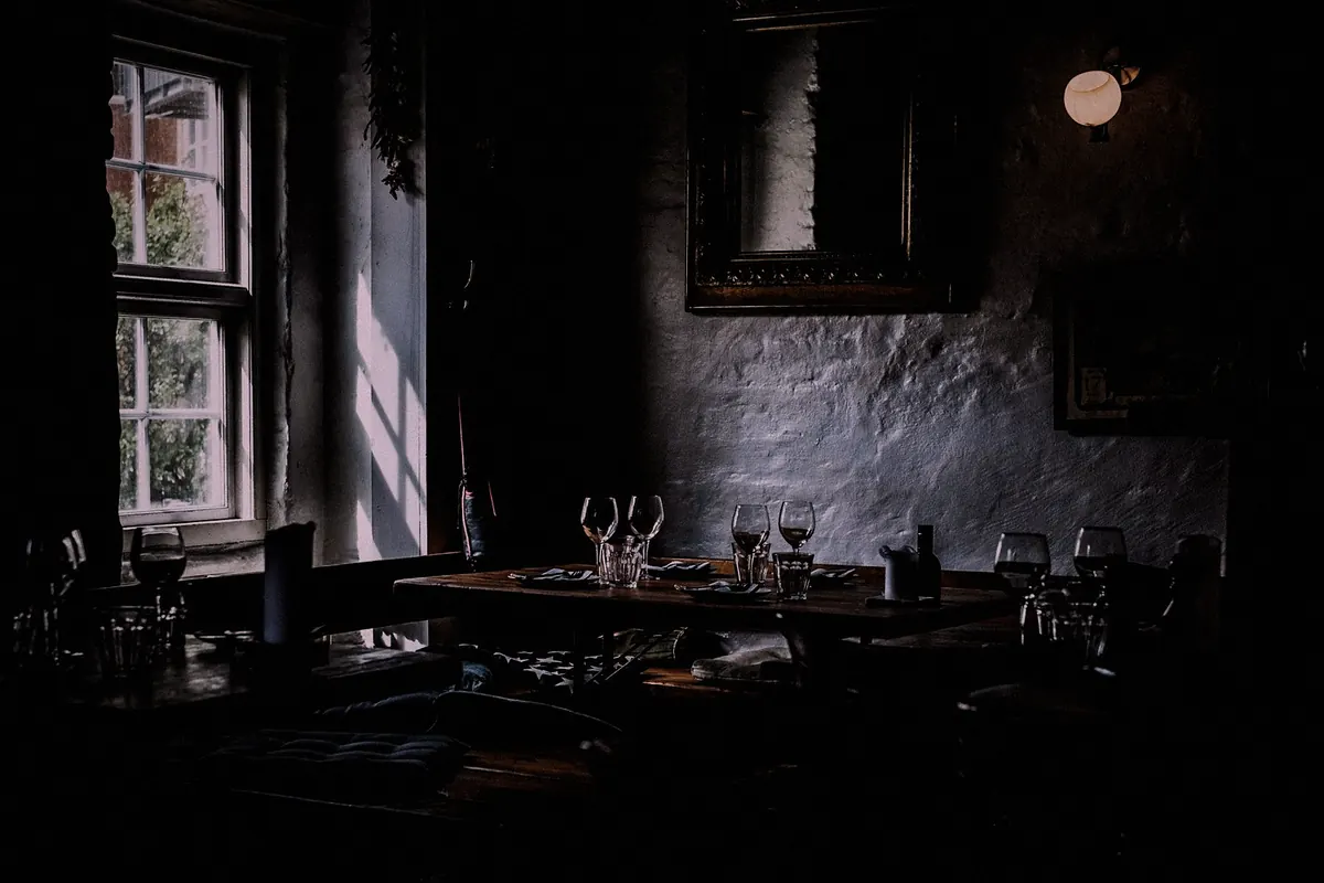 A dining room with stone walls in low light, one window letting in daylight