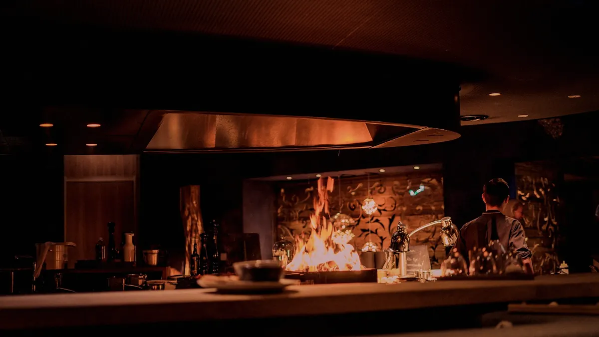 An open kitchen at night with a fire burning behind the counter under a steel hood