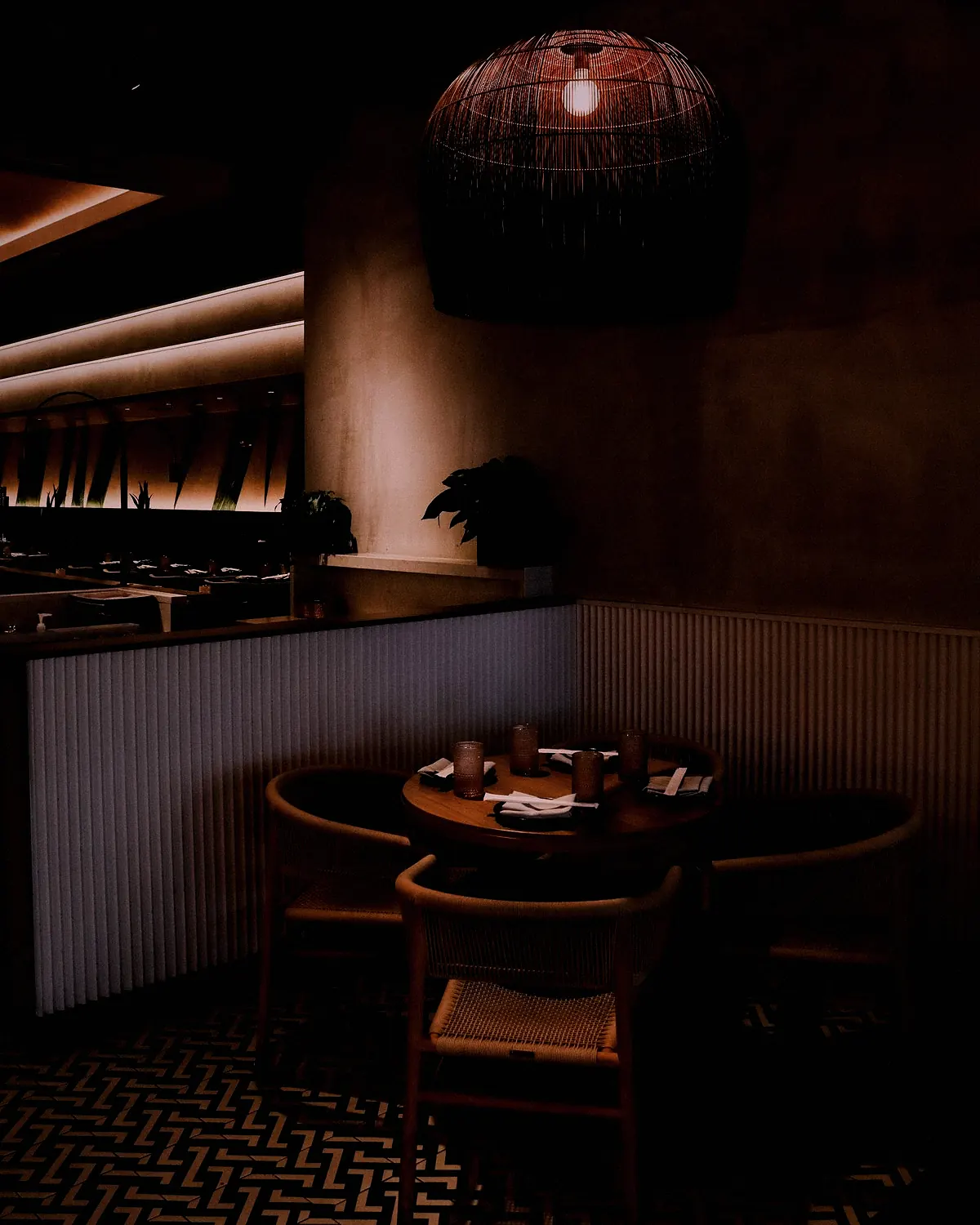 A round table for six under a woven lamp in a dark corner of the restaurant