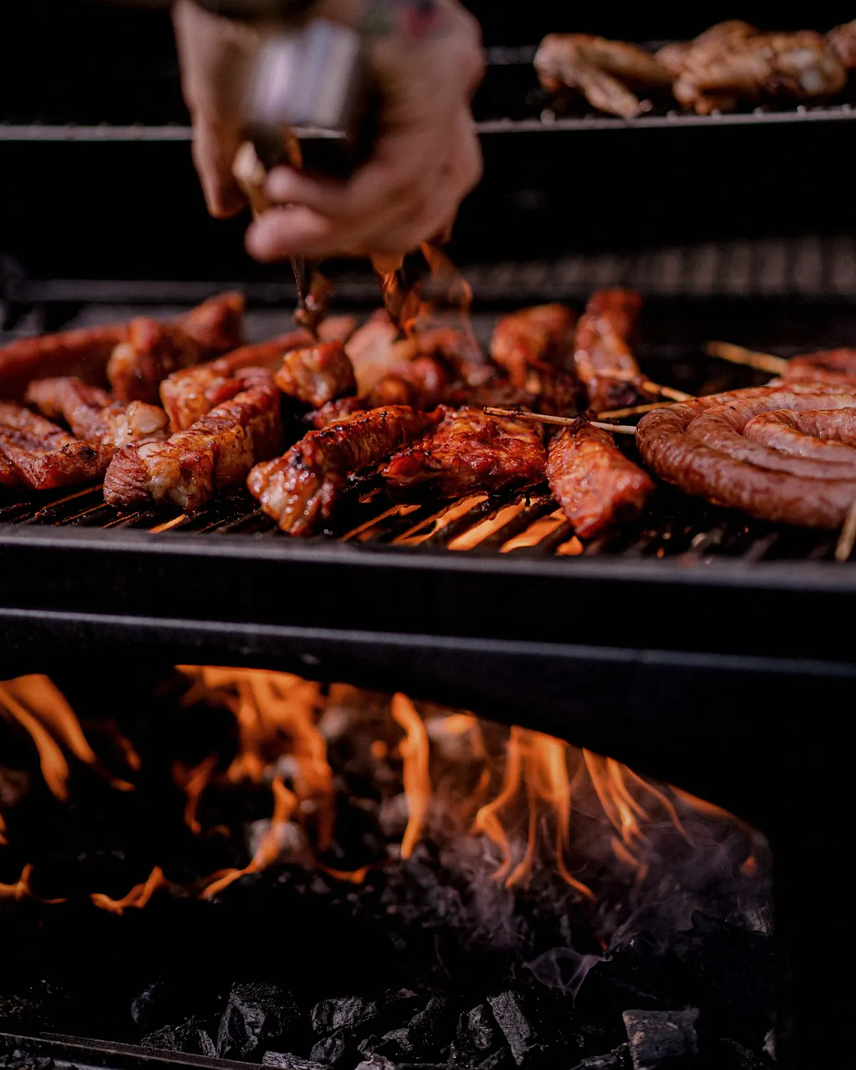 A cook turning skewers of meat on the grill, with the wood fire burning underneath