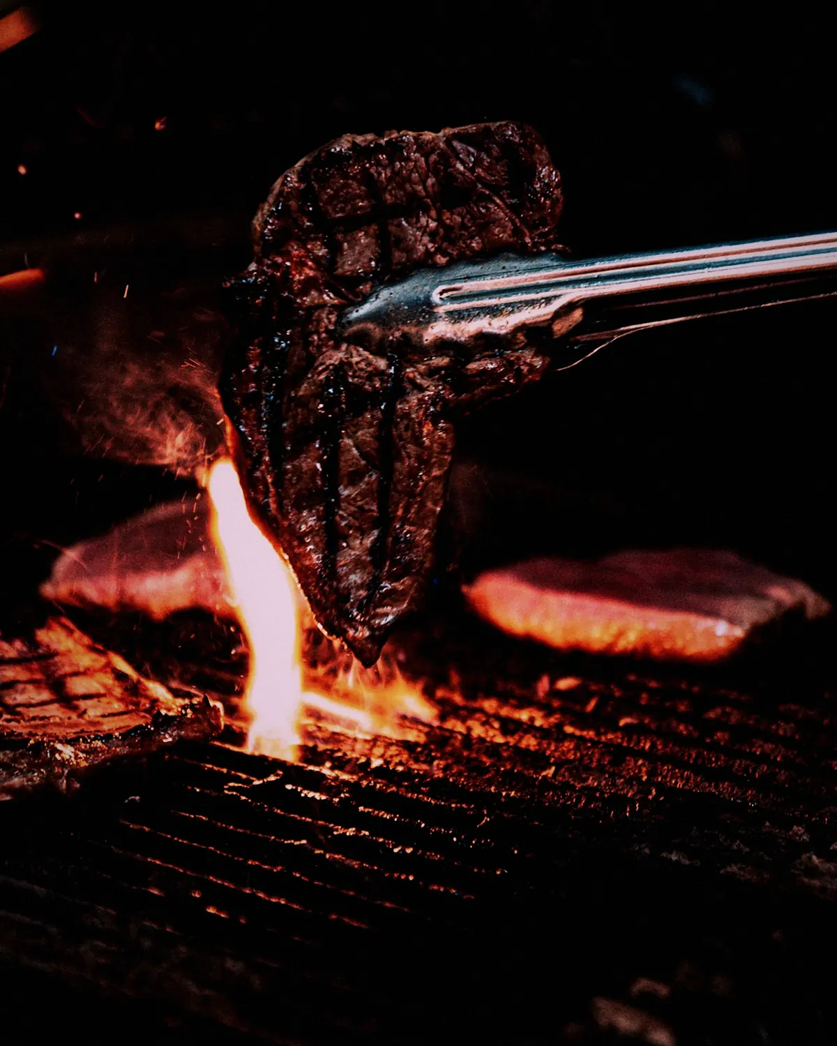 A steak lifted with tongs off a grill, flames below