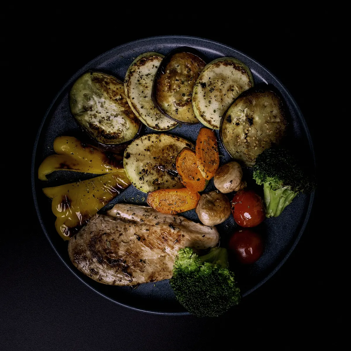 Charred courgette, peppers, aubergine and tomatoes on a dark plate, seen from above