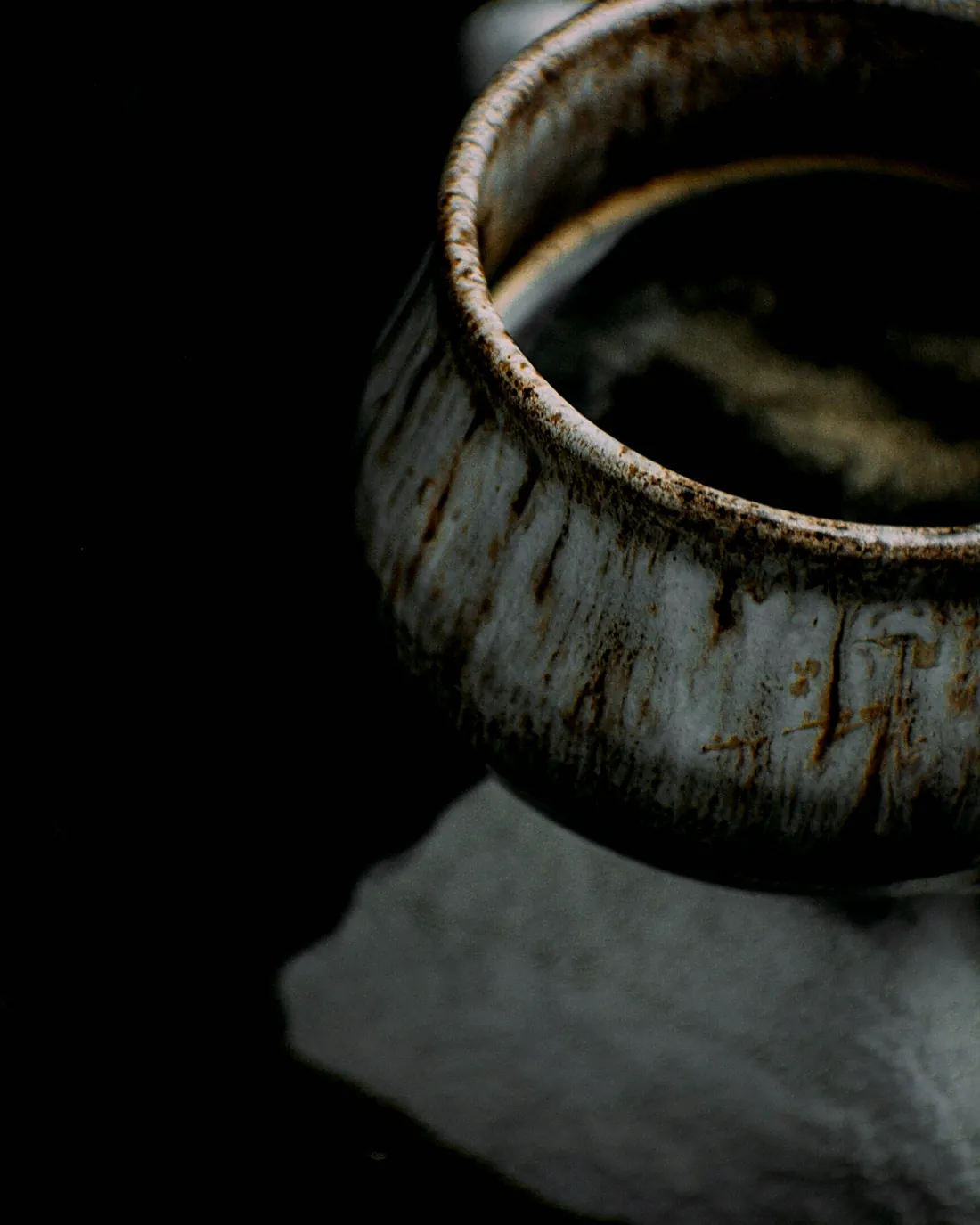 A small cup with a rust and grey glaze resting on a pale stone
