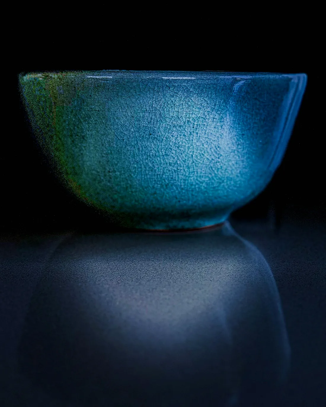 A teal glazed bowl on a dark table, its reflection below it