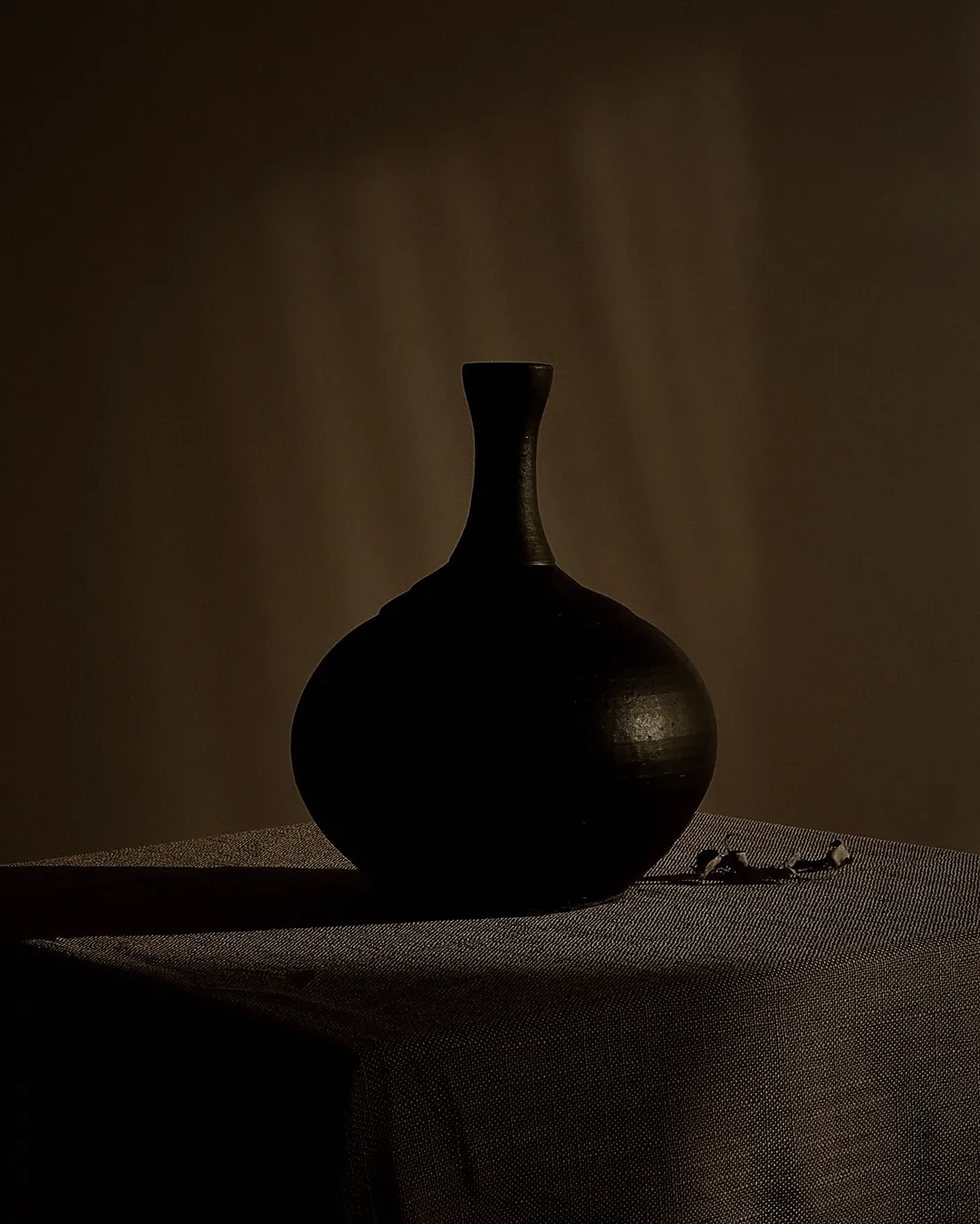 A round black vase with a narrow neck on a cloth covered table in low light