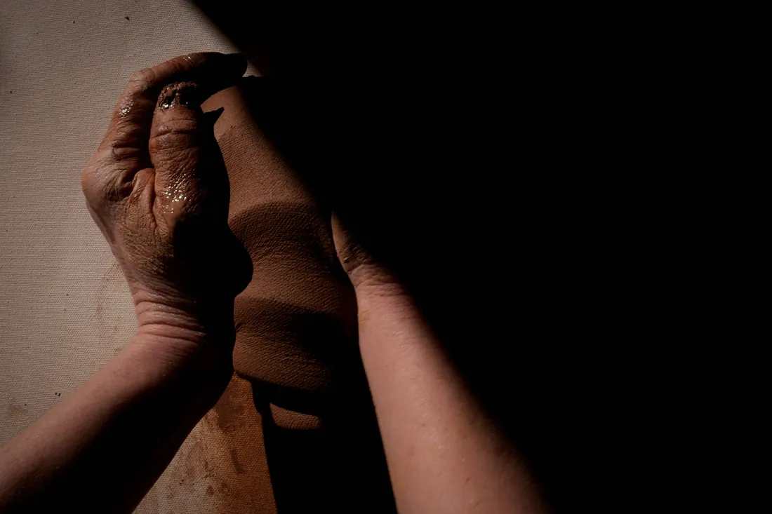 Two hands holding a column of wet clay, lit from the left against a dark wall
