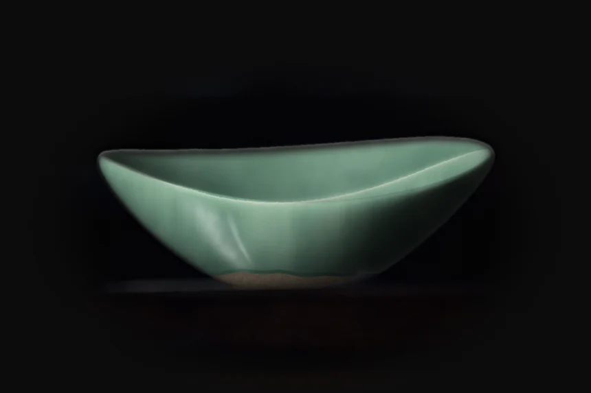 Oval stoneware bowl in celadon glaze, lit from the left on a dark shelf