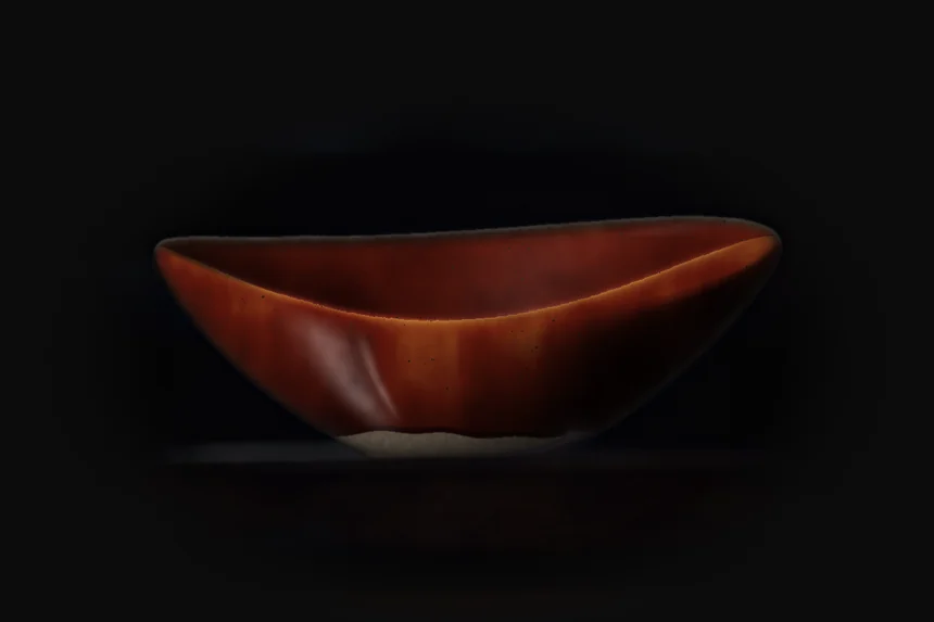 Oval stoneware bowl in oxide red glaze, lit from the left on a dark shelf