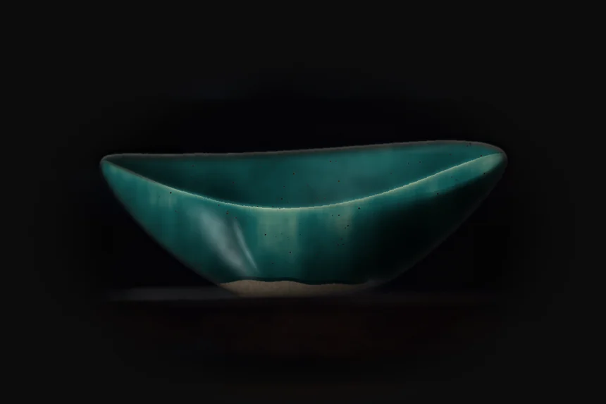 Oval stoneware bowl in copper teal glaze, lit from the left on a dark shelf