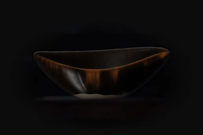Oval stoneware bowl in tenmoku glaze, lit from the left on a dark shelf