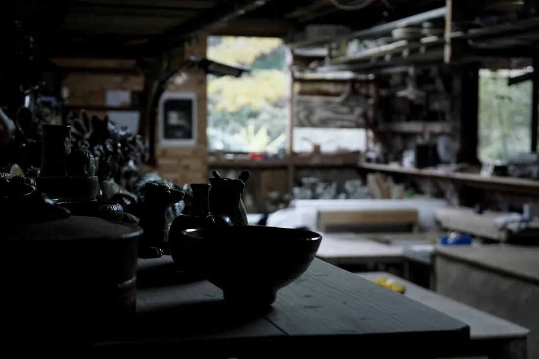 The studio in low light, with a dark bowl on a workbench and shelves of pots behind