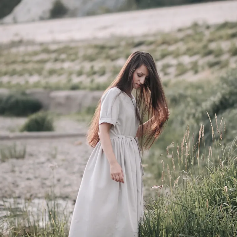 A woman in a long oat linen dress with short sleeves walks through tall grass by a river