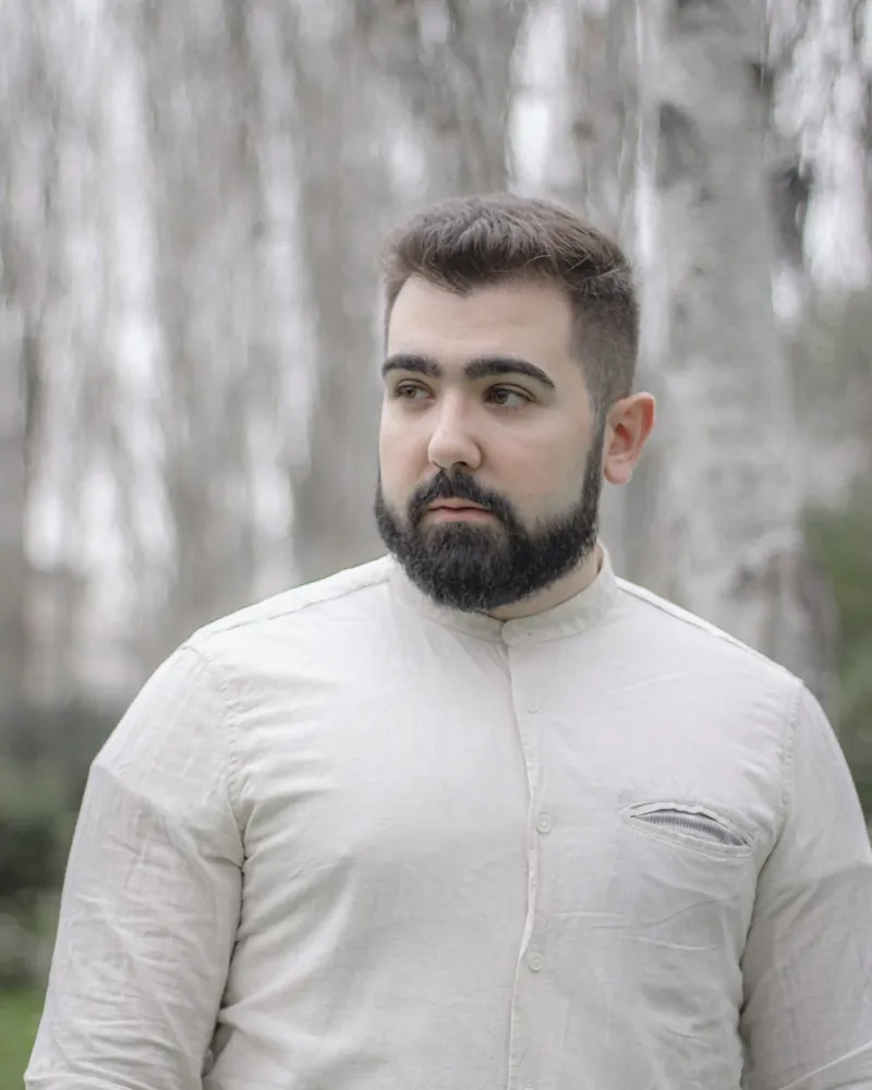 A bearded man in a white long sleeve linen shirt with a band collar and a chest pocket, birch trees behind him