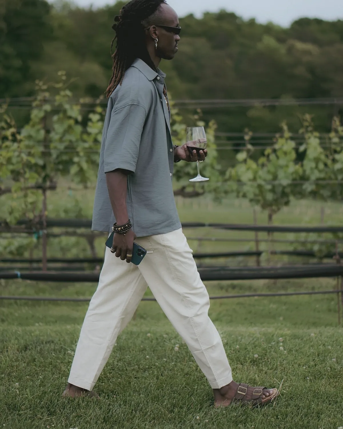 A man in a slate blue camp collar shirt and pale trousers walks past a vineyard holding a glass of red wine