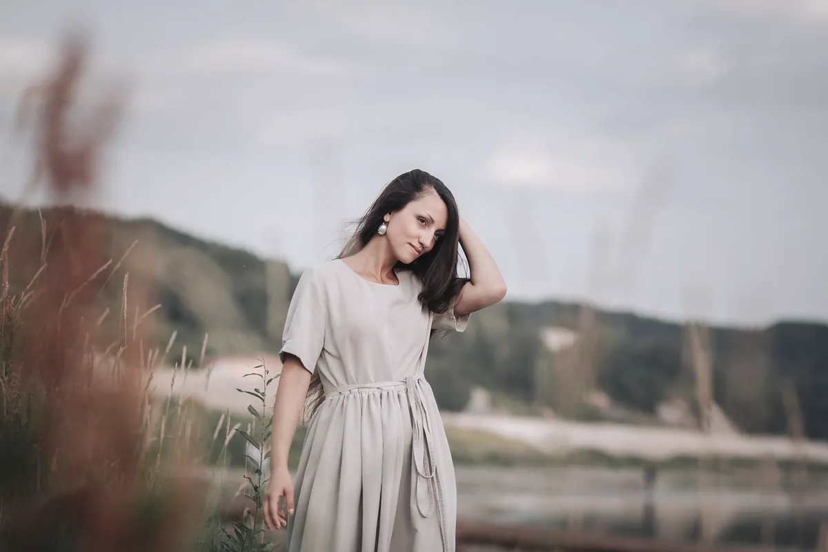 The same woman in the oat linen dress, a hand in her hair, with a river and hills behind her