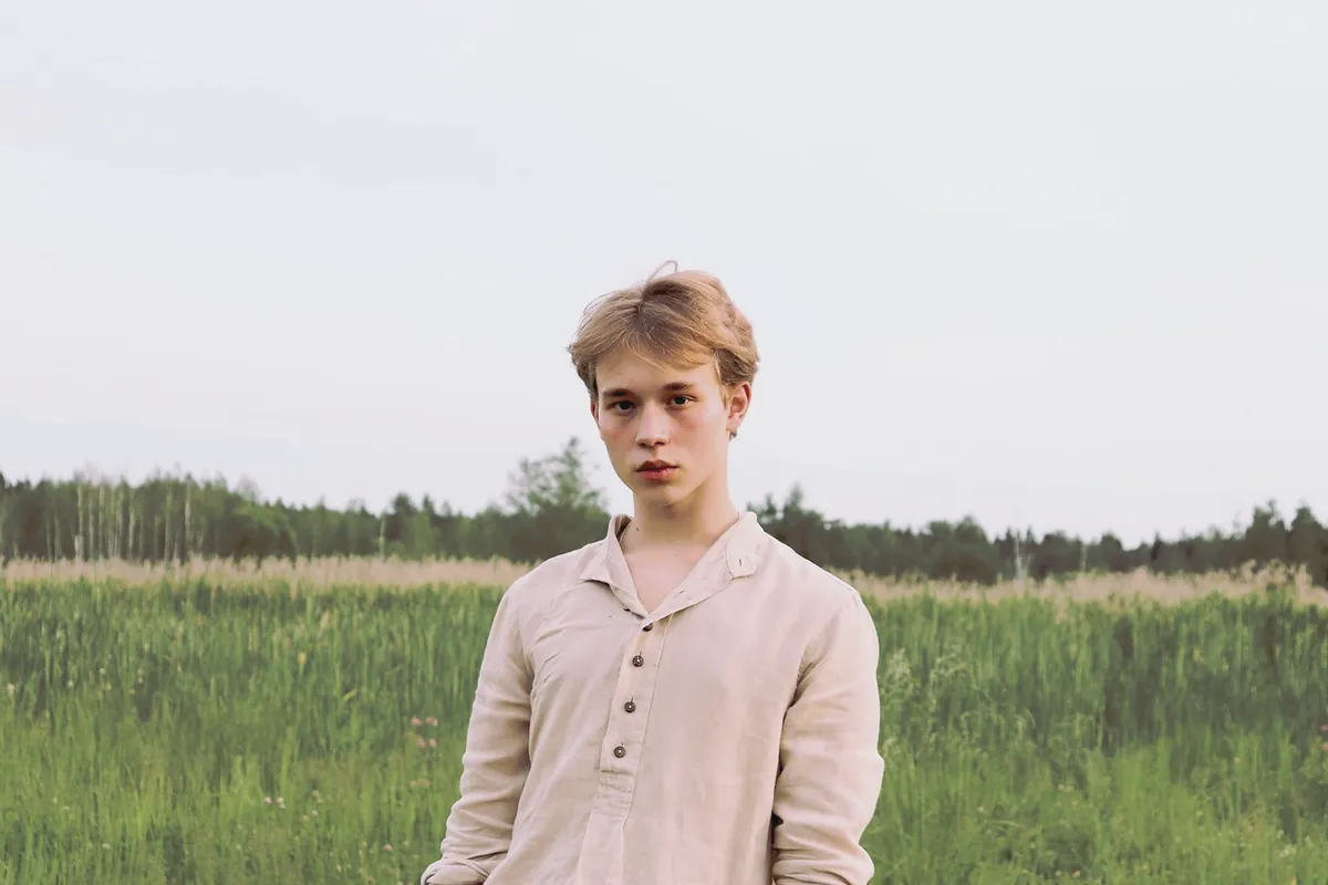 The same young man in the oat popover shirt in a wide green meadow under a grey sky