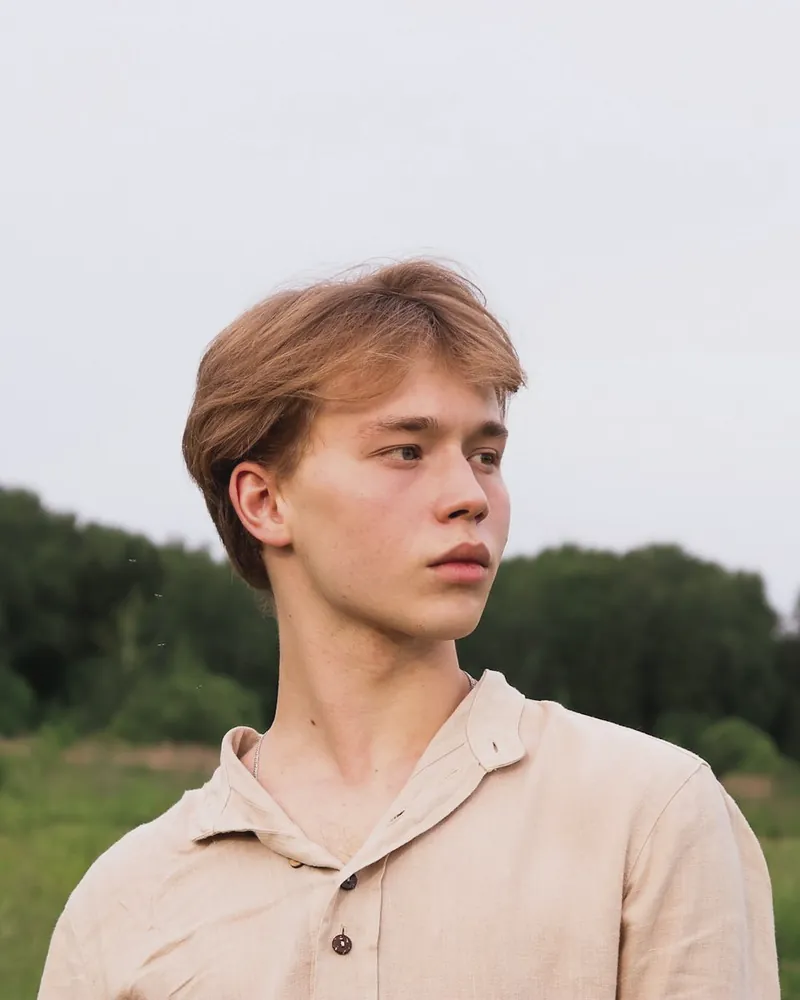 A young man in an oat linen popover shirt with an open collar stands in a meadow, trees behind him