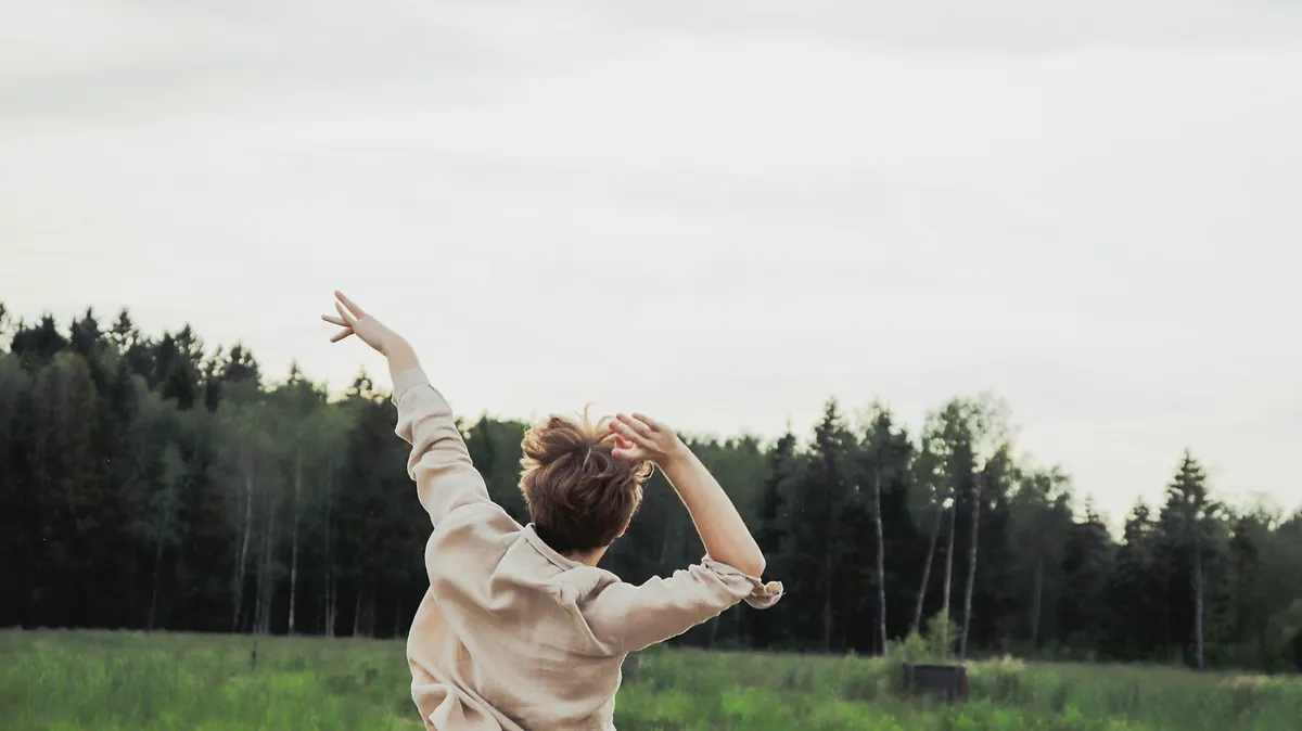 Seen from behind, someone in the oat popover shirt raises both arms in a meadow in front of a pine forest
