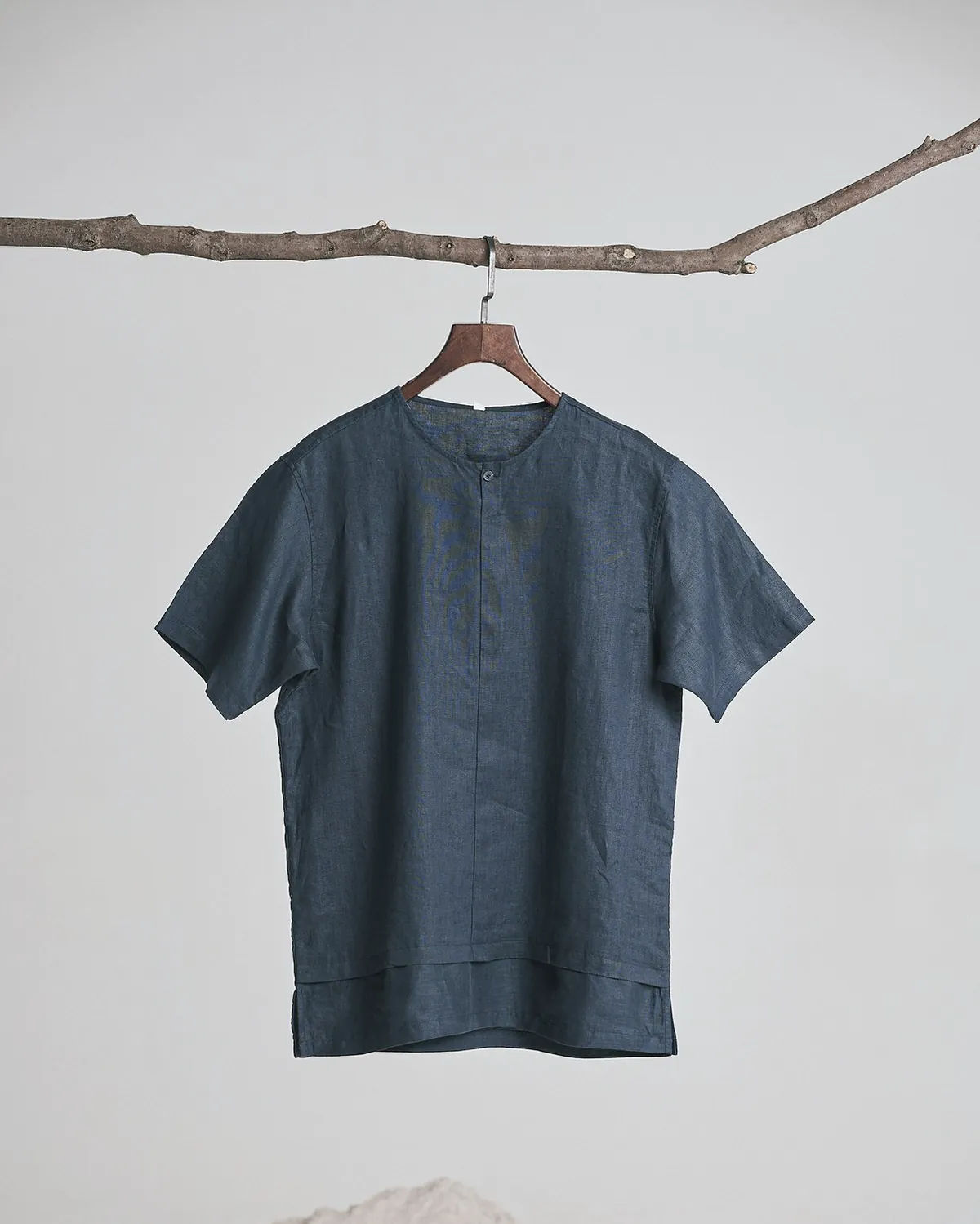 The same short sleeve band collar shirt in navy linen on a wooden hanger hung from a branch
