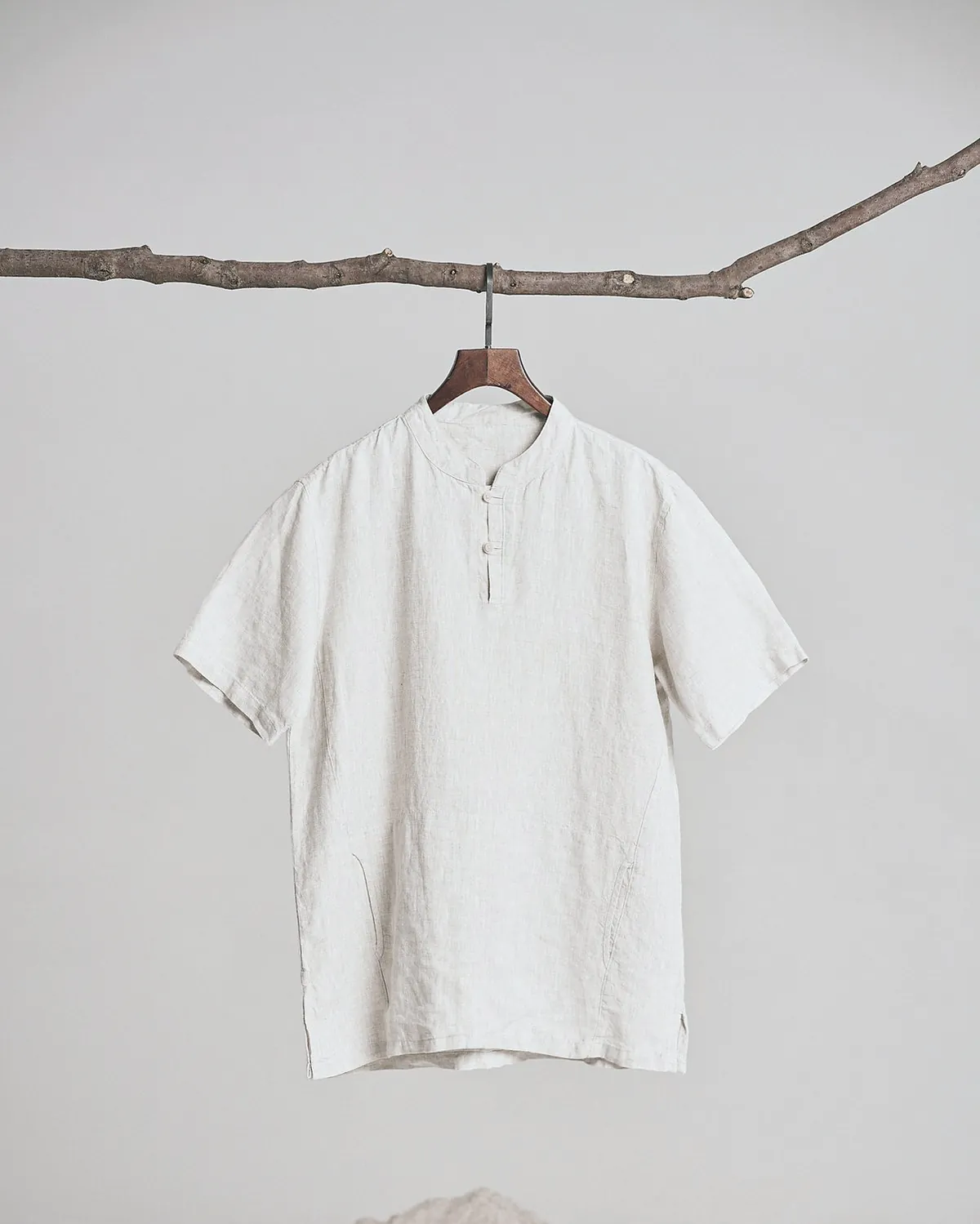 A short sleeve oat linen shirt with a band collar on a wooden hanger hung from a branch