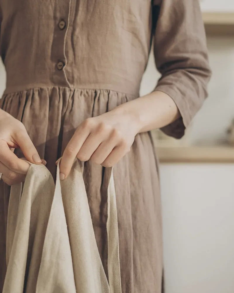 Close view of a walnut brown linen dress with a button front and a gathered skirt, hands holding an oat linen cloth