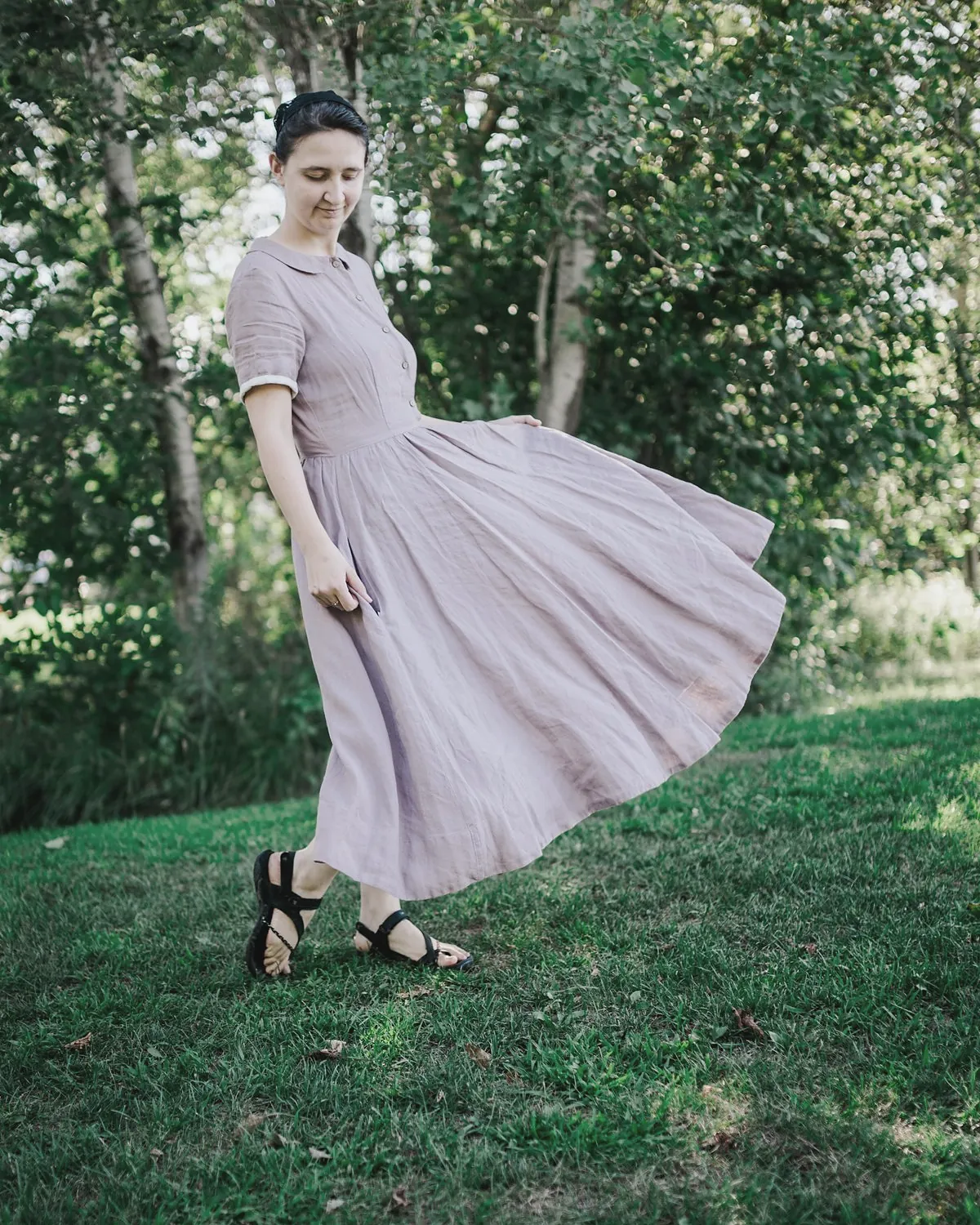 A woman in a lavender linen dress with a small round collar turns on a lawn so the full skirt swings out