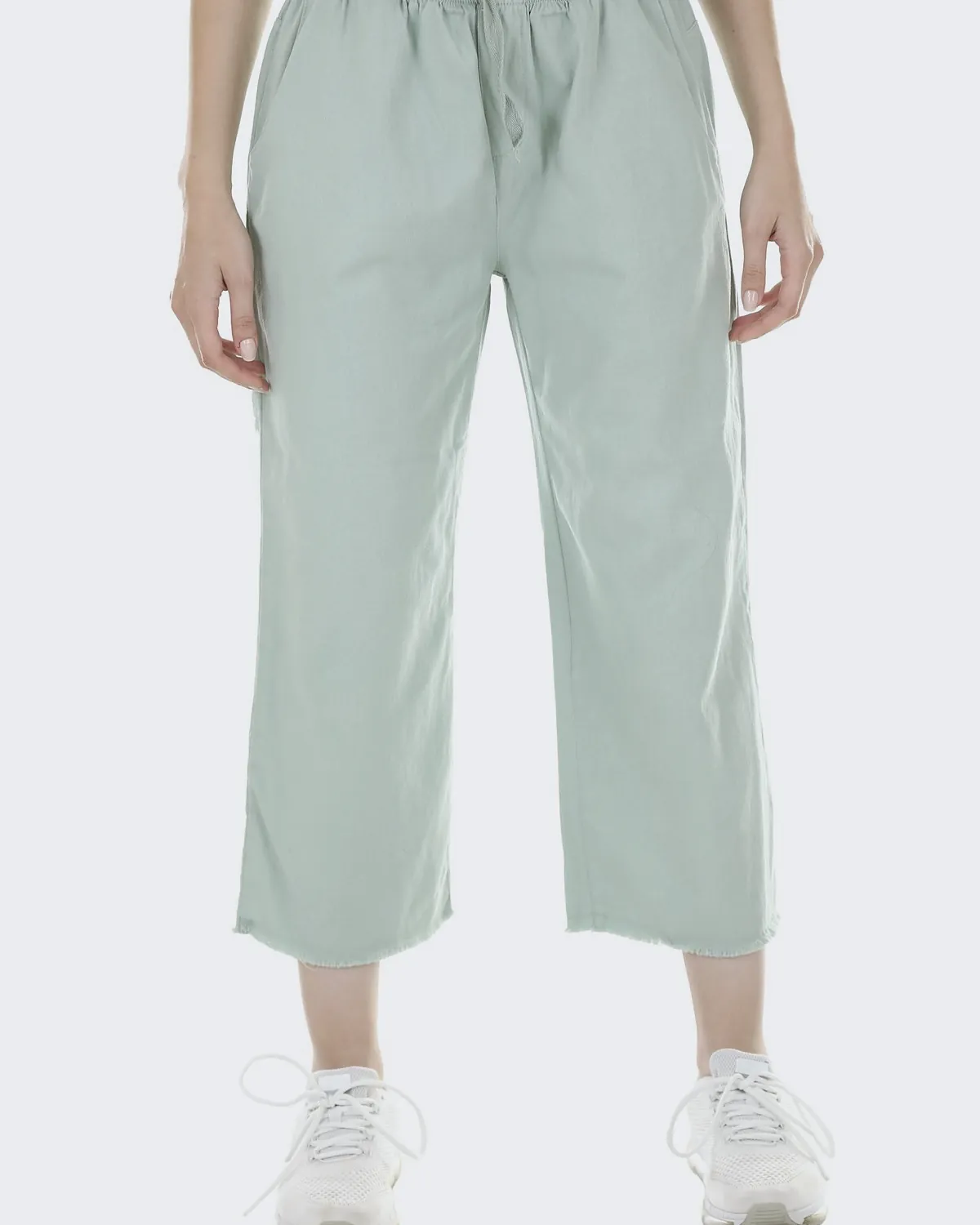 Sage green cropped linen trousers with a drawstring and a raw hem, worn with white trainers in a white studio